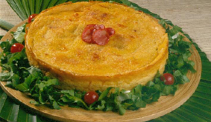Fotografia em tons de amarelo em uma bancada de madeira clara, uma toalha verde em formato de folha, uma tábua de madeira redonda com a torta caipira em cima dela e decorada com salada verde e tomates-cereja.