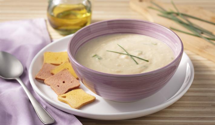Fotografia em tons de roxo e marrom de uma bancada de madeira com um prato raso branco, sobre ele um recipiente redondo roxo com o creme de palmito. Ao lado um paninho roxo com um colher. Ao fundo uma jarra com azeite e uma tábua de madeira com cebolinha.