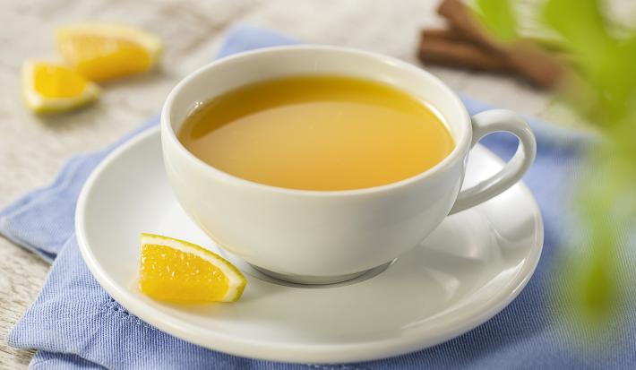 Fotografia em tons de azul, cinza e amarelo de uma bancada cinza com um paninho azul e sobre ele um prato branco redondo com uma xícara com chá de laranja. Ao fundo paus de canela e pedaços de laranja.