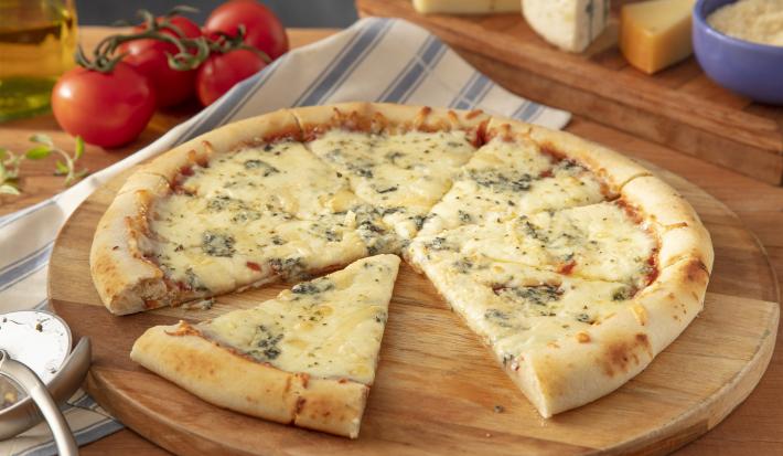Imagem de uma pizza redonda de 4 queijos com uma fatia deslocada. A fundo uma tábua de queijos e tomates.