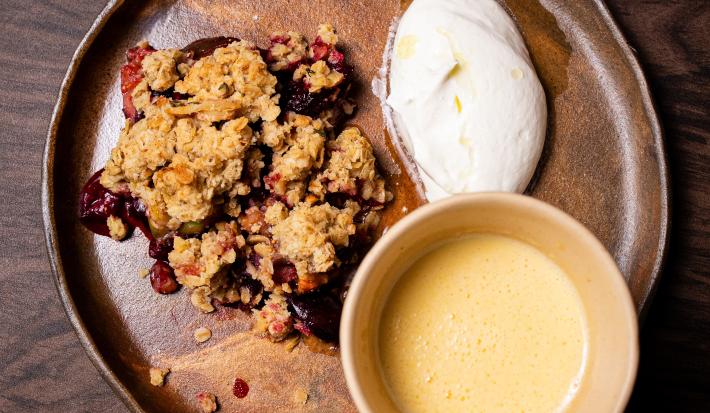 Fotografia vista de cima de um prato raso de vidro marrom, com um crumble de de frutas com creme inglês em um recipiente e chantilly de iogurte ao lado. O prato está sobre uma mesa de madeira.