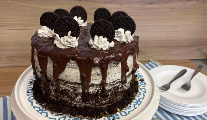 Fotografia em tons de branco, marrom, preto e azulado com um prato posicionado ao centro. Dentro do prato existe um bolo em formato redondo com recheio branco, cobertura de chocolate e enfeitado com chantilly e biscoitos.