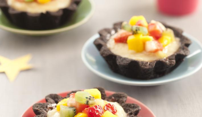 Fotografia em tons de vermelho em uma bancada de madeira clara com três pratinhos coloridos, vermelho, azul e verde e a tortinha de chocolate com frutas em cima dos pratinhos. Ao fundo, um copo rosa e um copo amarelo.