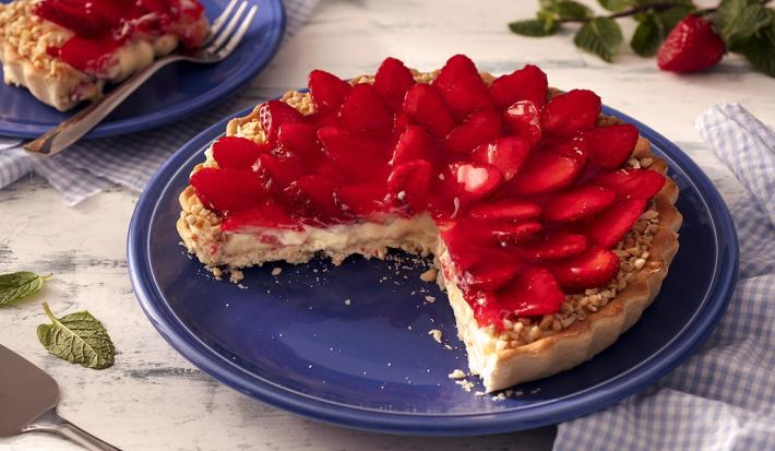 Fotografia em tons de vermelho em uma bancada de madeira de cor branca. Ao centro, um prato azul contendo a torta. Ao lado, há um pano azul quadriculado e ao fundo, um pires contendo uma fatia da torta.