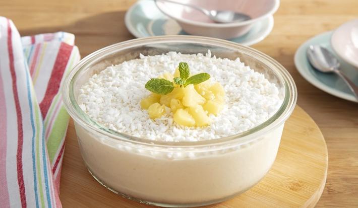 Foto em tons de branco da receita de gelado de abacaxi servida em um recipiente de vidro redondo sobre uma tábua circular de madeira com um pano colorido ao lado e xícaras brancas ao fundo