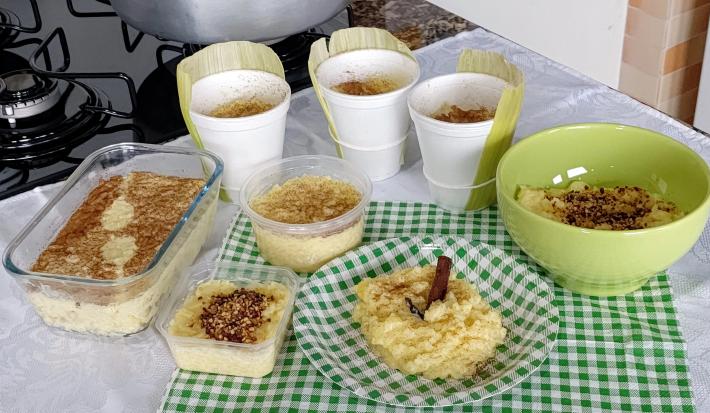 Fotografia de vários recipientes diferentes, sejam eles refratário de vidro, recipiente de vidro verde, prato colorido e copos grande de papel. Todos os recipientes têm dentro arros doce feito com milho verde e polvilhado com canela.