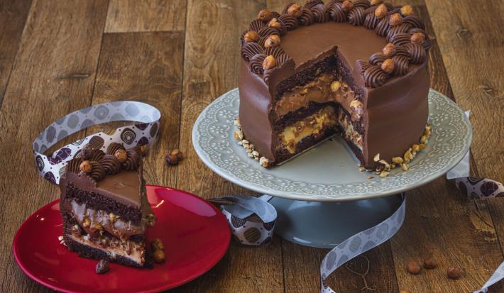 Foto da receita de bolo de chocolate com especiarias, sobre um prato cinza e sem uma fatia que está num prato vermelho do lado esquerdo. Tudo está em uma bancada de madeira decorada com uma fita de páscoa e pedaços de avelãs