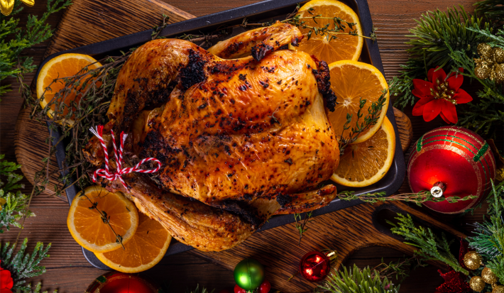 A imagem mostra um frango inteiro assado, decorado com fatias de laranja. A imagem de fundo remete ao Natal, com bolas vermelhas e festão
