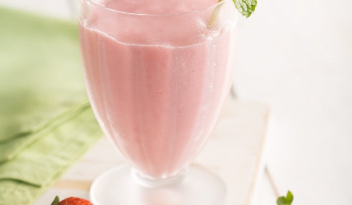 A imagem mostra um copo de smoothie rosa de morango, decorado com um raminho de hortelã fresca. O copo é transparente e tem uma base fosca, colocado sobre uma superfície de madeira clara. Ao redor, há três morangos frescos