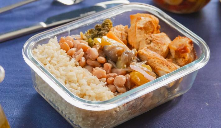 Marmita em pote de vidro transparente, com arroz integral, feijão, cubos de frango dourados e legumes assados coloridos (brócolis, cenoura, pimentões). Tudo bem distribuído, destacando cores vibrantes e textura fresca.