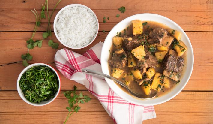 Fotografia em tons de marrom, branco, amarelo e verde de uma bancada marrom vista de cima. Contém um recipiente redondo branco com pedaços de carne e mandioca. Ao lado um pano branco e vermelho e dois potes redondos, um com arroz e o outro tempero