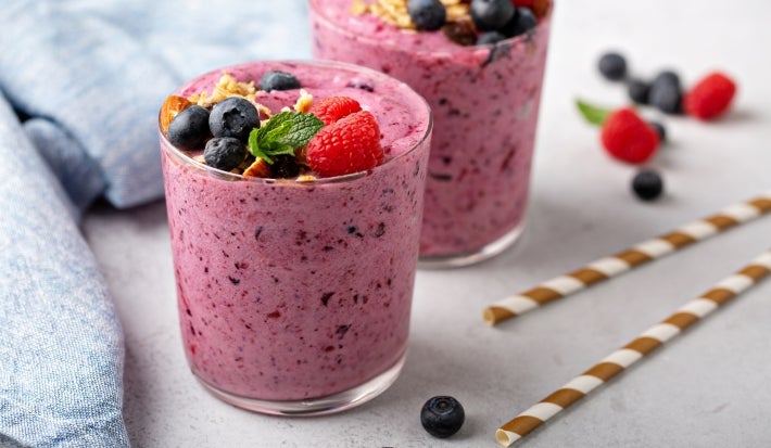 Fotografia em tons claros com dois copos de vidro ao centro. Dentro dos dois copos existe uma receita de smoothie de frutas vermelhas de cor rosa arroxeada. Por cima do smoothie, existe várias frutas vermelhas diversas.