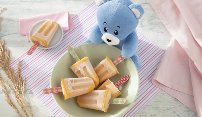 Foto da receita de Picolézinho de Manga com Banana. Observa-se um potinho verde claro com 5 picolés laranjas dentro, com cabinhos de madeira decorados. O pote está sobre um paninho listrado colorido.