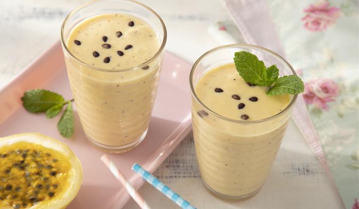 Receita de Suco Cremoso de Maracujá e Leite Moça. Observa-se dois copos com o suco decorados com hortelã e sementes de maracujá. Canudos coloridos rosa e azul e um maracujá cortado ao meio decoram a foto à esquerda.