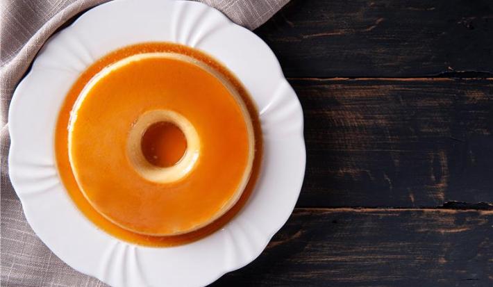 A foto descreve um pudim em um prato branco em uma mesa de madeira