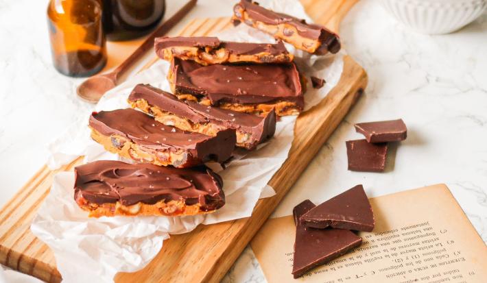 Fotografia em tons de marrom em uma bancada de madeira clara, uma tábua de madeira, barras de chocolate com pasta de amendoim ao centro e pedaços de chocolate soltos ao lado.