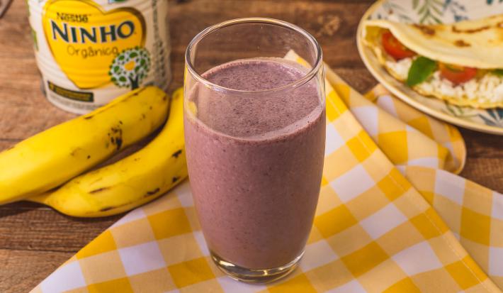 Fotografia em tons de amarelo de uma bancada com um pano xadrez amarelo e branco e um copo com a bebida de açaí. Ao fundo bananas, uma lata de NINHO e um prato com uma crepioca.