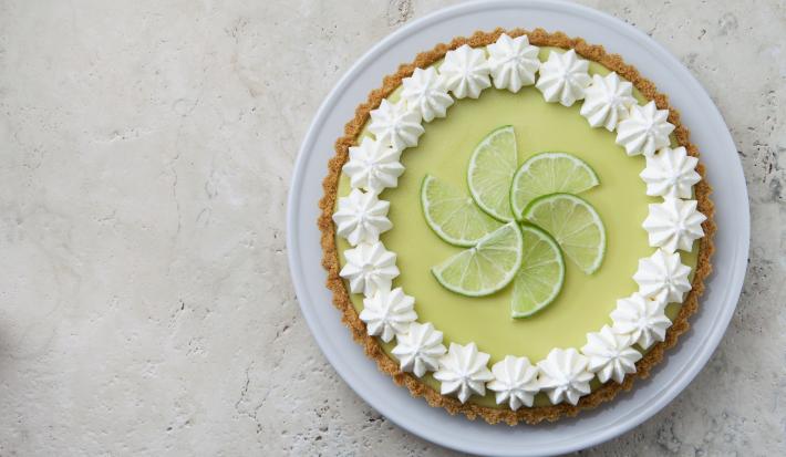 Foto de uma torta limão em cima de um prato branco e com fatias de limão decorando o centro da torta.