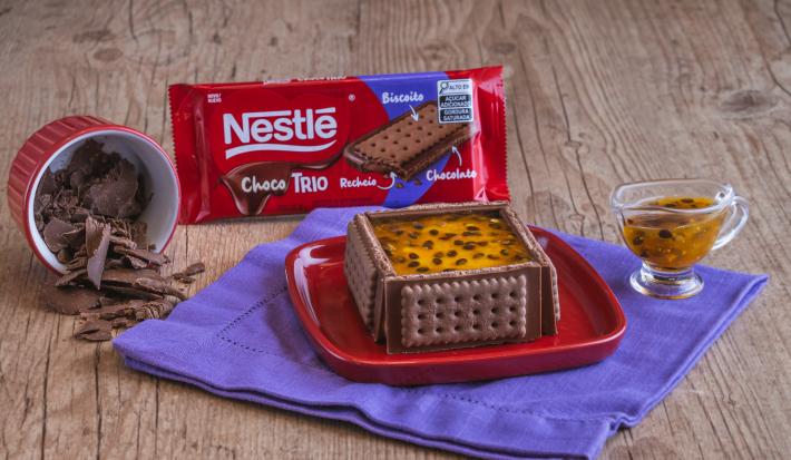 Fotografia de um bolo mousse de maracujá sobre um prato quadrado vermelho. Ao redor do bolo nota-se o chocolate choco trio, além de uma calda de maracujá por cima do bolo.
