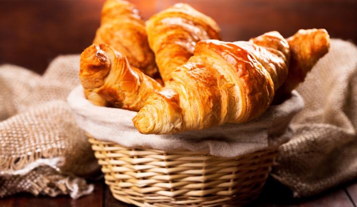 Fotografia de cestinhas de palha com um pano de tom bege. Dentro da cestinha no centro da foto, tem vários pães tipo croissant.