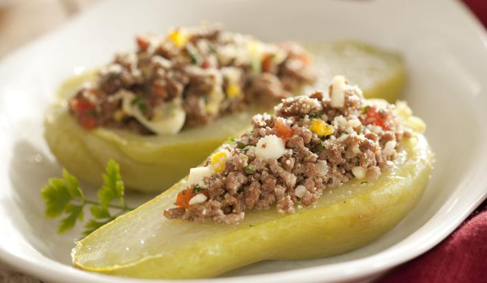 Fotografia em tons de branco, verde e vinho, ao centro refratário branco com duas metades de chuchu com carne moída, ao lado guardanapo vinho, pote branco ao fundo, tudo sobre bancada de madeira envelhecida.