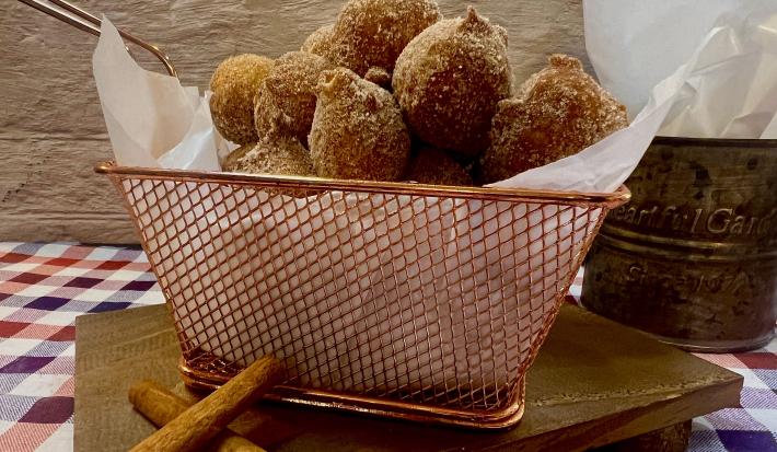 Foto da receita de Bolinho de Chuva. Observa-se uma cesta de metal com guardanapo e os bolinhos de chuva dentro sobre uma tábua de madeira em cima de uma toalha quadriculada.