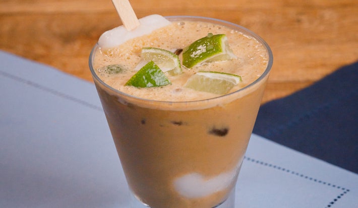 Copo baixo com bebida cremosa e cubos de limão no topo, apoiado sobre tecido claro e mesa de madeira.