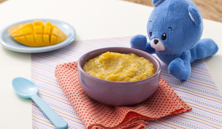 Fotografia em tons de amarelo e azul de uma bancada de madeira com um paninho listrado, um potinho roxo com o creminho de manga, ao lado uma colher azul e um ursinho de pelúcia azul. Ao fundo, um pratinho azul com um pedaço de manga cortada.