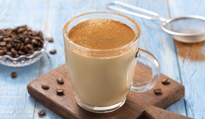 Foto da receita de Cappuccino Quente Cremoso. Observa-se uma caneca de vidro transparente com o cappuccino polvilhado de canela em cima de uma pequena tábua de madeira sobre uma superfície de madeira azul clara. Ao fundo, grãos de café e uma peneira.