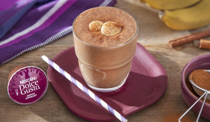 Foto da receita de iced coffee com banana e cacau servida em um copo largo de vidro sobre um prato vinho com um canudo listrado, em cima de uma mesa de madeira com uma banana e um paninho roxo ao fundo