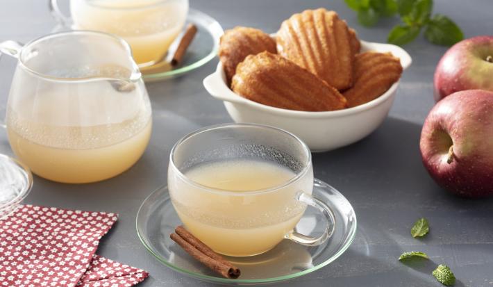 Imagem de uma xícara de chá de maçã, com um pedaço de canela no pires ambientado com maçãs e um pote de madeleines