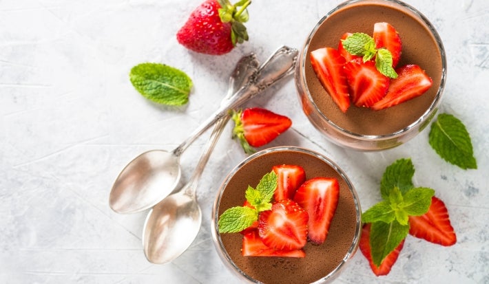 Fotografia em tons claros e vista de ciam de dois copos decorativos de tamanho normal com a sobremesa de taça moranguda, finalizada com chocolate e decoração de morangos em pedaços e folhas de hortelã.
