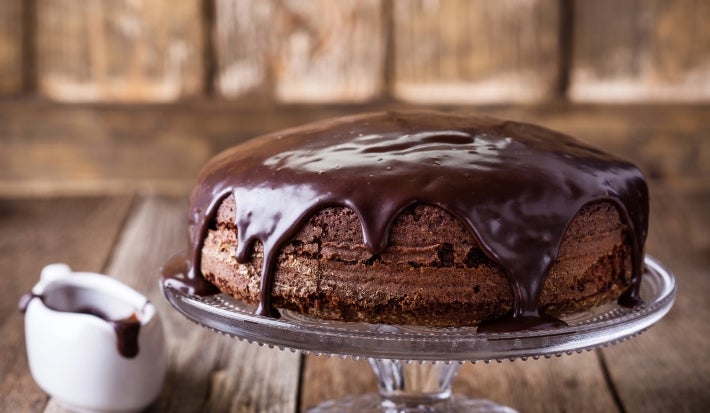 Fotografia de um bolo de chocolate em um suporte de vidro, coberto com ganache escura que escorre pelas laterais. Ao lado, um pequeno jarro de cerâmica com ganache, em um cenário acolhedor de madeira.