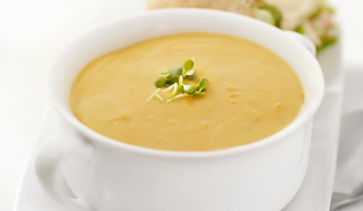 Fotografia em tons claros, de um recipiente de vidro na cor branca sobre um prato retangular branco. Dentro do recipiente, um caldo de mandioquinha, ao fundo, dois lanches pequenos, tudo sobre uma mesa na cor branca.
