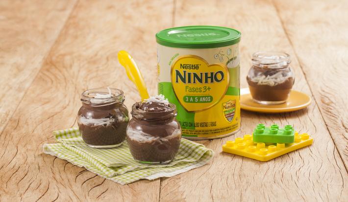 Fotografia em tons de verde e amarelo em uma bancada de madeira clara, um paninho verde xadrez com dois potinhos de acrílico com a sobremesa de prestígio dentro dele. Ao lado, brinquedinhos, uma lata de Ninho Fases e mais um potinho com a sobremesa.