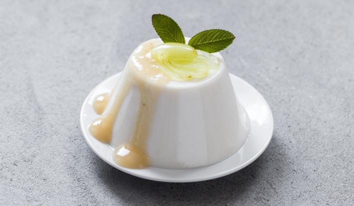 Fotografia de uma mousse de uva verde, com um creme e uma unidade de uva em cima, e para decorar, duas folhas de hortelã. A sobremesa está em um prato pequeno de sobremesa na cor branco, que está sobre uma mesa cinza. Fotografia de uma mousse de uva verde, com um creme e uma unidade de uva em cima, e para decorar, duas folhas de hortelã. A sobremesa está em um prato pequeno de sobremesa na cor branco, que está sobre uma mesa cinza.