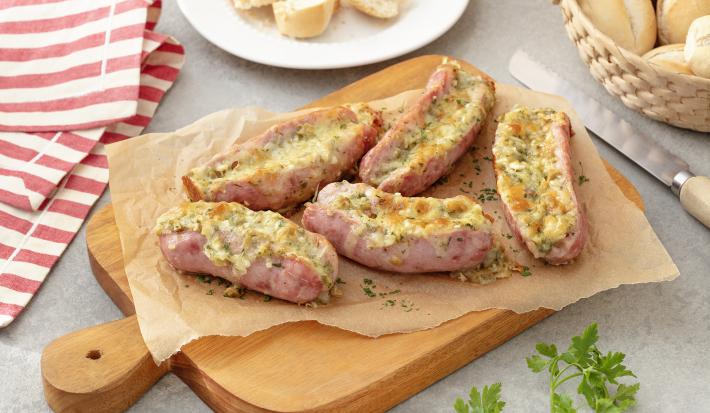 Foto em tons de bege e cor de rosa da receita de linguiça recheada servida em 5 porções sobre uma tábua de madeira com uma faca prateada ao lado. Ao fundo há um prato branco com fatias de pão francês e um pano listrado