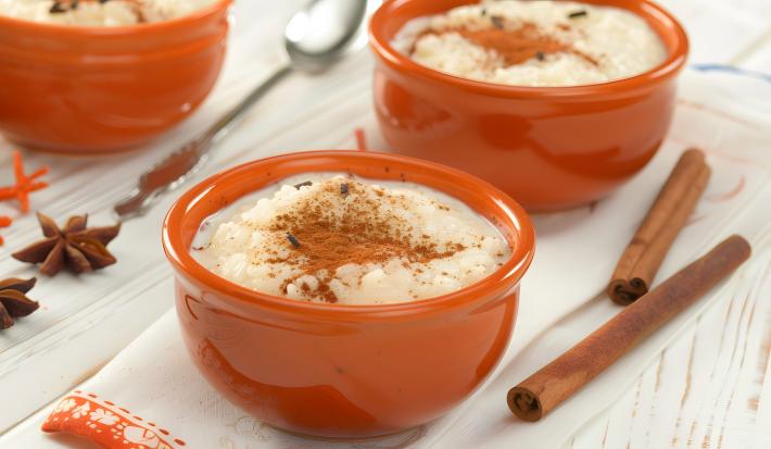 Fotografia em tons claros de três recipientes fundos e pequenos marrons sobre uma mesa clara com alguns paus de canela e cravos da índia. Dentro dos recipientes têm arroz doce com canela.