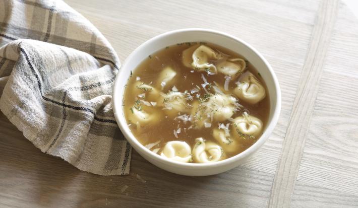 Fotografia de um recipiente branco fundo com uma sopa de capeletti, sobre uma mesa de madeira. Ao lado, um pano branco com linhas na cor cinza e preto. Fotografia de um recipiente branco fundo com uma sopa de capeletti, sobre uma mesa de madeira. Ao lado, um pano branco com linhas na cor cinza e preto.