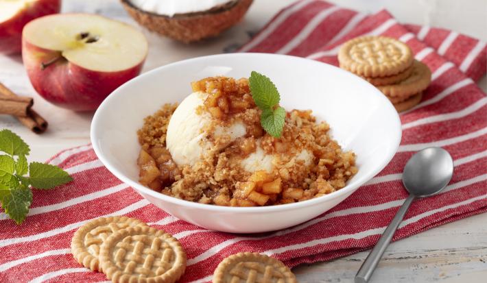 Fotografia em tons de branco e vermelho de uma bancada branca com um paninho listrado branco e vermelho, sobre ele um recipiente redondo branco com o sorvete. Ao lado uma colher e biscoitos tostines. Ao fundo uma maçã e um coco.