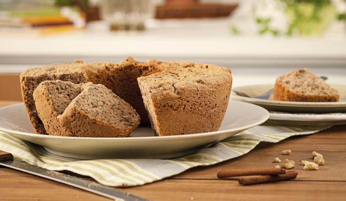 Fotografia em tons de marrom em uma mesa de madeira com um prato raso branco apoiado em um pano cinza listrado em bege e o bolo de maçã e nozes. Ao lado, um prato com um pedaço do bolo e paus de canela espalhadas pela mesa.