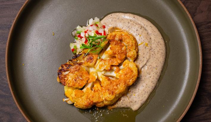 Fotografia em tons de marrom com um prato cinza ao centro. Em cima do prato existe uma receita de couve-flor tostada na panela com creme de castanhas e vinagrete