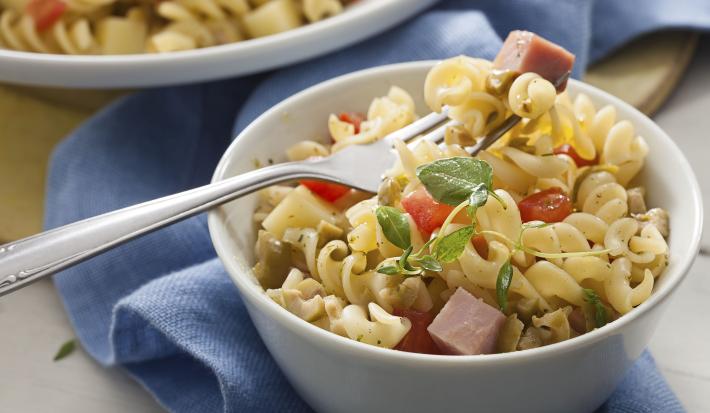 Fotografia em tons de azul em uma bancada de madeira com um pano azul ao fundo, um prato redondo fundo com a salada de macarrão tipo parafuso, com bacon e tomate.