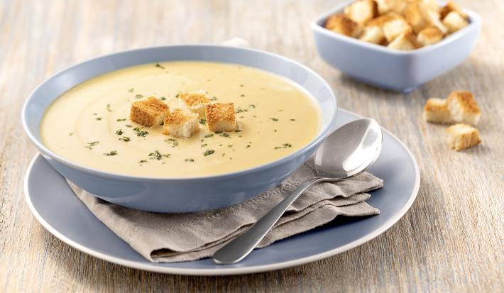 Foto da receita de Sopa de Mandioquinha com Baunilha em uma tigela azul clara, em cima de um prato azul claro e um guardanapo cinza, sobre uma tábua de madeira clara e com croutons em um pequeno pote de decoração.