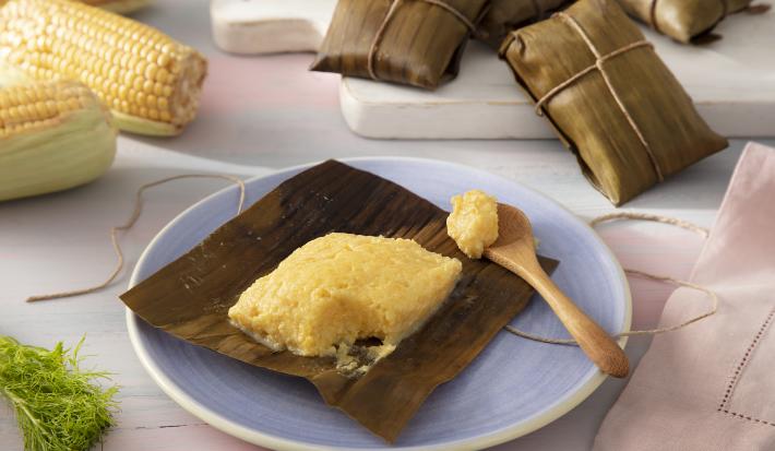Foto da receita de Pamonha de Leite Moça, Milho Verde e Erva Doce. Observa-se a pamonha aberta em um prato de sobremesa de cerâmica com uma colherinha ao lado direito. Atrás, tem milhos na espiga e pamonhas fechadas e embrulhadas para decorar.