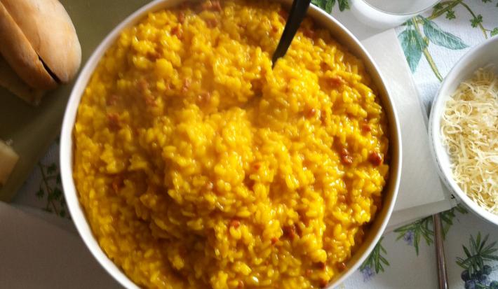 Fotografia vista de cima de um refratário fundo branco com arroz cremoso com açafrão. Ao lado direito, dois pratos e dois copos brancos, um bowl pequeno branco com queijo ralado, e no outro lado, um prato quadrado verde com alguns pães.