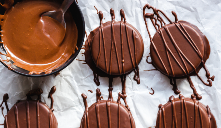 Imagem mostra 5 alfajores redondos cobertos por chocolate ao leite escuro, dispostos sobre um papel manteiga branco. Ao lado um recipiente com chocolate derretido e uma colher.