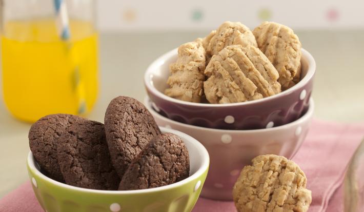 Fotografia em tons de rosa em uma bancada de madeira clara com potinhos verde, branco e marrom com bolinhas brancas em cima de um pano rosa e biscoitinhos de chocolate e aveia dentro dos potinhos.