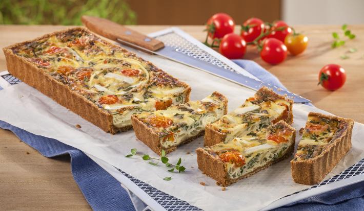 Fotografia em tons branco, dourado e vermelho, a direita e ao alto tomates-cereja e folhas de tomilho, ao centro travessa branca com quiche assada e fatiada, sobre papel manteiga e guardanapo azul em bancada de madeira.