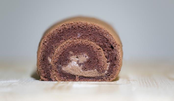Fotografia de um bolo de rolo de chocolate, em tons de marrom, ao centro de uma mesa de madeira clara.